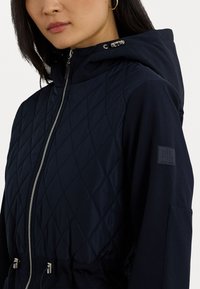 Giacca trapuntata blu navy con zip frontale, cappuccio con lacci e patch del logo sulla manica. Texture liscia e design elegante.