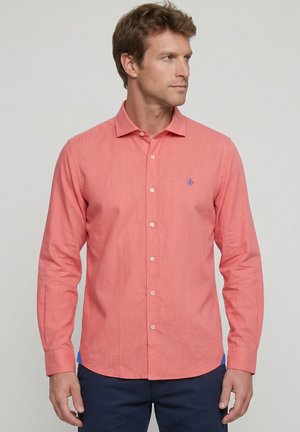 Hombre con camisa coral de manga larga abotonada con pequeño logo azul, mirando hacia la derecha, combinado con pantalones azul marino sobre fondo gris claro.