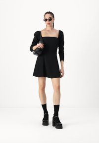 Vestido mini negro con mangas abullonadas y escote cuadrado, combinado con zapatos de plataforma negros gruesos y calcetines negros hasta el tobillo. Pequeña bolsa de hombro negra.