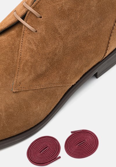 Bruine suède desertboot met tan leren veters, met een ronde neus en rubberen zool. Bevat twee bordeauxrode ronde veters naast de boot.