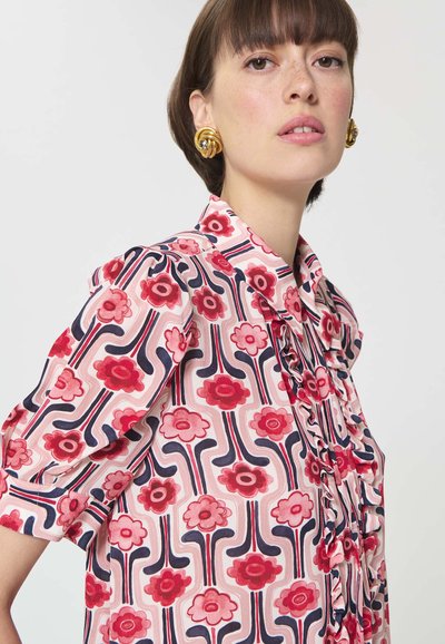 Femme aux cheveux courts bruns portant des boucles d'oreilles dorées en forme de spirale et un chemisier rose avec des motifs floraux rouges et des lignes ondulées, détail froncé à l'avant.