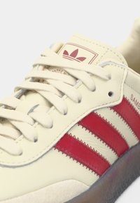 Αθλητικό παπούτσι από κρέμ δέρμα με τρεις κόκκινες ρίγες, επενδεδυμένο κολάρο και vintage ετικέτα με λογότυπο. Διαθέτει υφασμένα κορδόνια και καουτσούκ εξωτερική σόλα.