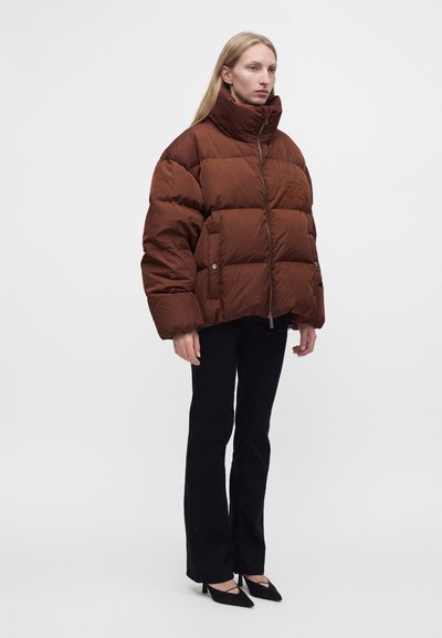 Femme portant une doudoune marron avec col montant, pantalon noir et talons pointus noirs, debout devant un fond blanc uni.
