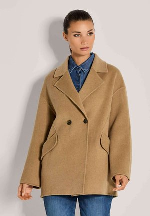 Vrouw in een beige dubbelzijdige wollen jas over een blauwe denim shirt en blauwe spijkerbroek, staand tegen een effen lichte achtergrond.