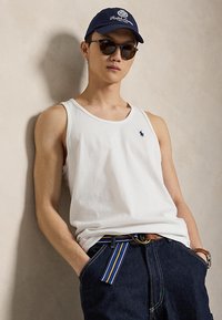Jeune homme portant un débardeur blanc, des lunettes de soleil foncées, une casquette marine et un jean foncé avec une ceinture rayée bleu-jaune, posant devant un mur beige uni.