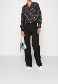 Pantalons noirs taille haute, coupe ample, associés à un chemisier noir à manches longues avec un motif abstrait blanc. Petit sac irisé et baskets blanches.