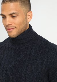 Suéter de punto cableado en azul marino con cuello alto, que presenta patrones texturizados y acentos en punto de canalé en el cuello y el dobladillo.