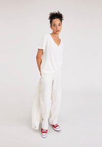 T-shirt bianco a collo a V, di taglio rilassato, abbinato a pantaloni bianchi a gamba larga e sneakers rosse in tela. Design semplice e pulito senza motivi visibili.
