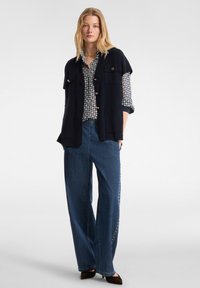 Frau mit gemusterter Bluse, navyblauer ärmelloser Jacke mit Knöpfen, weiten Jeans mit Seitennieten und braunen spitzen Schuhen, stehend vor einfarbigem Hintergrund.