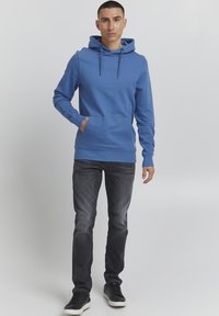 Sudadera de algodón azul con un bolsillo tipo canguro, capucha con cordón y dobladillo acanalado. Llevada con jeans oscuros y zapatillas negras.