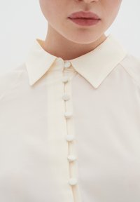 Cremefarvet skjorte med klassisk krave og en knapstolpe med runde, hvide knapper. Stoffet ser ribbet ud med en blød tekstur.