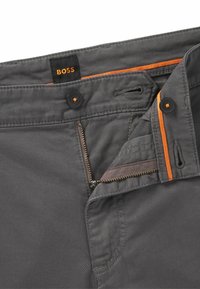 Pantaloni grigi con chiusura a bottone e cerniera, caratterizzati da dettagli arancioni lungo la vita e i bordi delle tasche. Dettaglio di tessuto testurizzato.