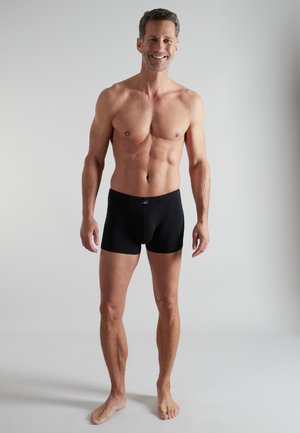 Smiling fit man standing barefoot wearing black boxer briefs against a plain light gray background.