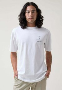 Camiseta de algodón blanca con mangas cortas. El diseño frontal incluye un gráfico de ancla y ola en gris claro. Corte relajado.