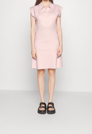 Kobieta ubrana w bladoróżową, bezrękawnikową sukienkę z kołnierzykiem oraz czarne platformowe sandały z dwoma klamrami, stoi na tle jednolitego białego tła.