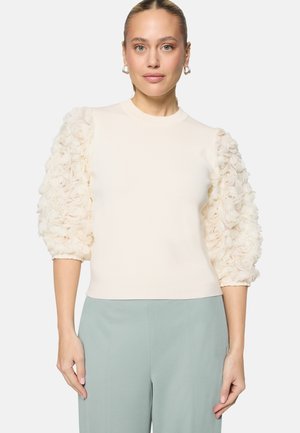 Femme portant un pull crème avec des manches bouffantes florales texturées et un pantalon taille haute bleu clair, debout devant un fond uni.