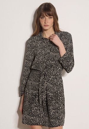 Woman wearing a black long-sleeve dress with white irregular dots, standing against a plain light background.