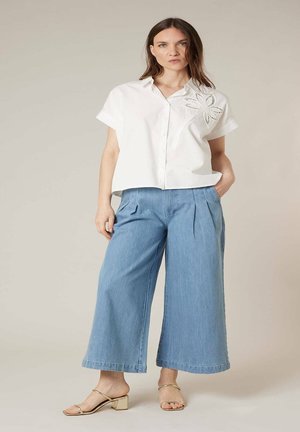 Femme debout, main dans la poche, portant une chemise blanche à manches courtes avec un détail floral et un pantalon en denim bleu à jambes larges.