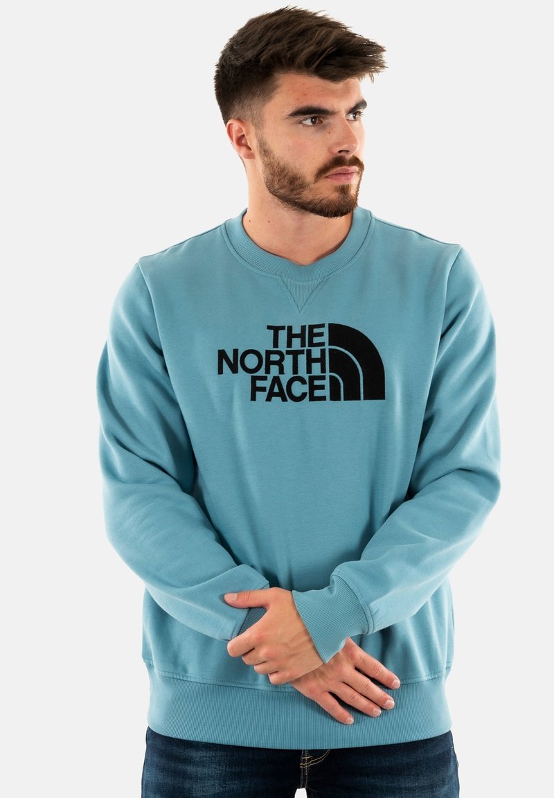 Felpa leggera azzurra con logo nero, polsini a costine e scollatura tonda. Il modello è in piedi con le mani incrociate sulla vita.