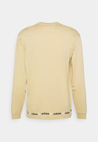 Långärmad beige t-shirt i bomull med rund halsringning och svarta accenter som har det upprepade vita "adidas"-logotypen på fållen.