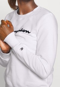 Person som bär en vit sweatshirt med svart "Champion"-logotyp över bröstet och en liten logotyp på vänster ärm, händerna knäppta nära bröstet.