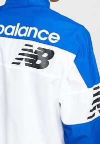 Blå och vit jacka med hög krage, utrustad med en framträdande svart "NB"-logotyp och texten "balance" över ryggen. Lättviktigt tyg.