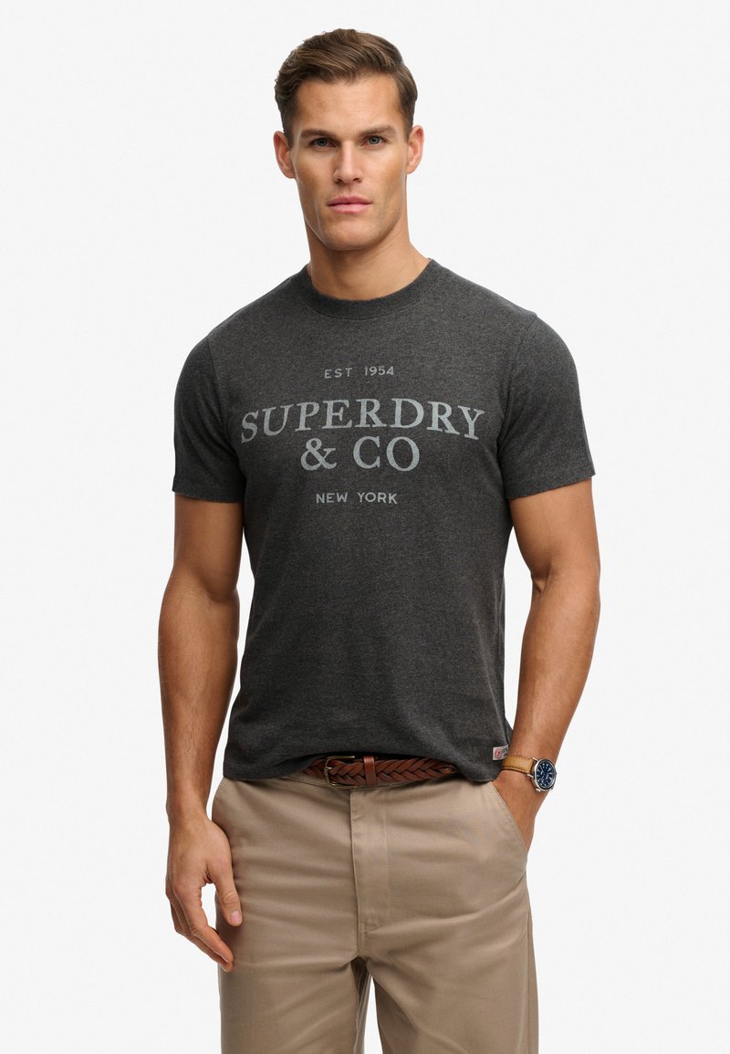 Graues kurzärmeliges T-Shirt mit rundem Ausschnitt und dem Aufdruck "SUPERDRY & CO" in hellgrau auf der Vorderseite, kombiniert mit beigen Hosen.