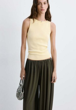 Femme portant un haut sans manches jaune pâle et un pantalon ample vert foncé, tenant un petit sac à main à motif gris sur un fond uni.