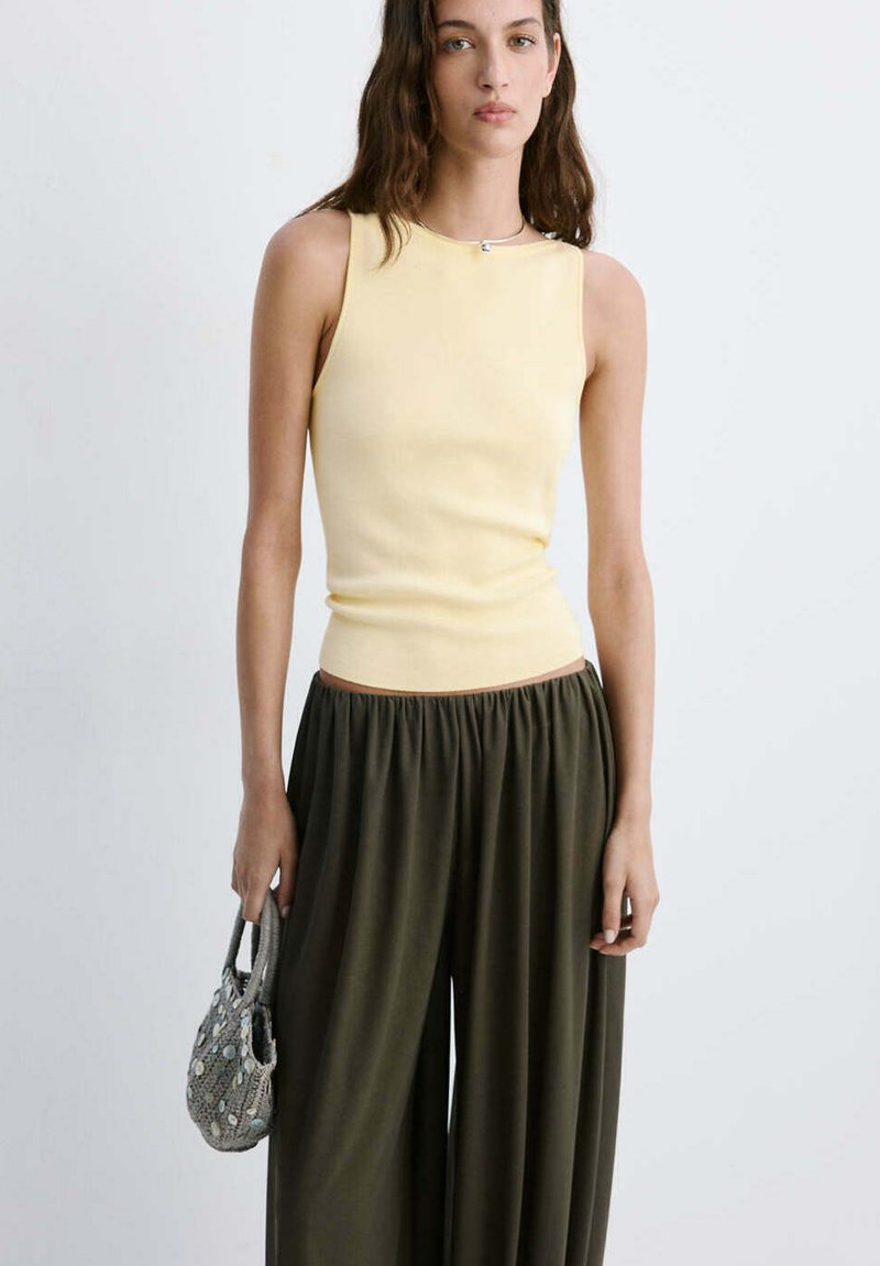 Femme portant un haut sans manches jaune pâle et un pantalon ample vert foncé, tenant un petit sac à main à motif gris sur un fond uni.