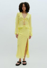 Geel doorschijnende blouse met ruffles en een trektouw, gecombineerd met een bijpassende rok met een split aan de zijkant. G gedragen met zwarte platte sandaal.