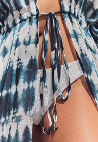 Niebiesko-biała narzutka tie-dye z rozciętym wzorem i zapięciami na wiązania, w zestawie z białymi, marszczonymi majtkami bikini z teksturowanymi detalami.