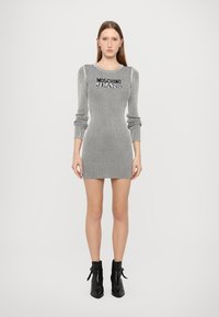 Šedé proužkované šaty s dlouhým rukávem a přiléhavým střihem. Na prsou je černě vytištěno "Moschino Jeans". Nosené s černými kotníkovými botami.
