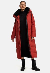 Longue doudoune rouge avec une capuche détachable en fourrure noire, fermeture éclair et accents noirs. Portée par-dessus une robe tricotée noire et des bottes noires.