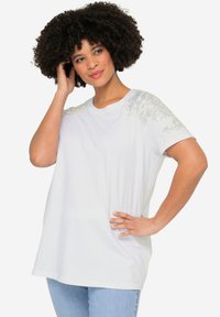 Weißes T-Shirt aus weichem Stoff mit dekorativen silbernen Verzierungen an den Schultern. Kurze Ärmel und lockere Passform.