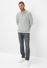 Man die tegen een witte achtergrond staat, gekleed in een grijze poloshirt met lange mouwen, donkere jeans en witte sneakers, met zijn handen achter zijn rug.