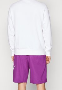 Person som bär en enkel vit sweatshirt och lila träningsshorts, stående med ryggen vänd mot betraktaren.