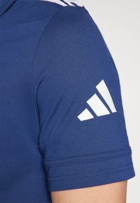 Sininen urheilupaita lyhyillä hihoilla, jossa on valkoinen kolmoisraita-design olkapäässä ja valkoinen logo hihassa. Silkkinen pinta.