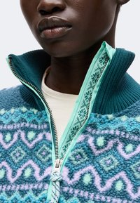 Gros plan d'une personne portant une veste polaire bleue et turquoise à motifs, avec une fermeture éclair partielle et un col côtelé, par-dessus une chemise blanche.