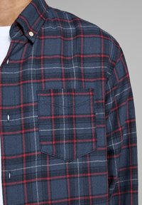 Tmavě modrá a červená károvaná flanelová košile s knoflíkovým límečkem, s náprsní kapsou a bílým prošíváním na texturované látce.