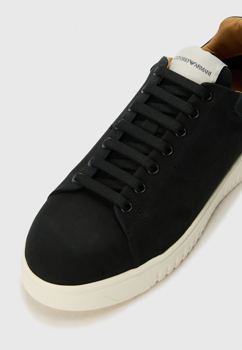 Baskets noires avec un dessus en cuir lisse, des lacets texturés et une semelle en caoutchouc blanche. Présente un logo sur la languette et un design minimaliste.