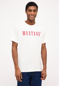 Balta medvilnė marškinėliai su žodžiu "MUSTANG", atspausdintu ryškiai raudonomis raidėmis per krūtinę. Apvalus iškirpimas ir trumpomis rankovėmis.