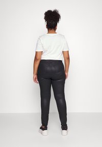 Kvinna med lockigt hår i en vit t-shirt, svarta skinny jeans och svart-vita sneakers som står vänd mot en vit vägg.