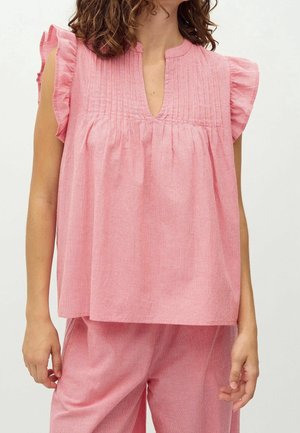 Femme portant un haut rose ample sans manches avec des épaules à volants et un pantalon assorti, debout devant un fond uni.