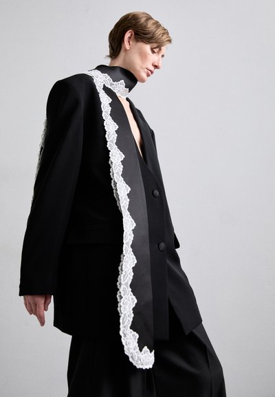 Zwarte oversized blazer met een diepe V-hals, voorzien van witte kant langs de kraag en de voorranden, uitgevoerd in een soepel materiaal.
