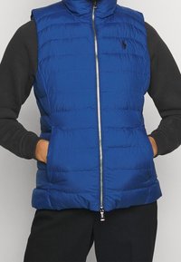 Chaleco de plumas azul con un alto cuello, costuras horizontales, cierre frontal con cremallera y dos bolsillos laterales. El material parece suave y ligero.