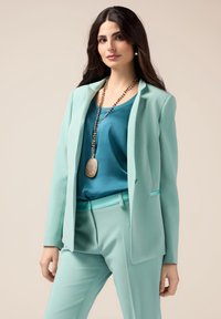 Mintgrüner maßgeschneiderter Anzug mit einem einreihigen Blazer, passender Hose und einer tief tealfarbenen Bluse. Enthält eine lange Anhängerkette.