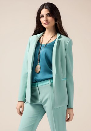 Completo sartorial verde menta con blazer a un bottone, pantaloni abbinati e una blusa verde petrolio. Presenta una lunga collana con pendente.