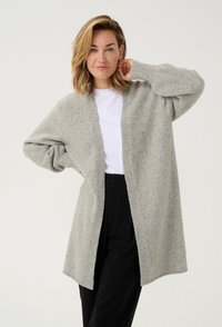 Hellgraue Strickjacke aus weichem, strukturiertem Material. Mit offenem Vorderteil, langen Ärmeln und lockerer Passform. Kombiniert mit einem weißen Oberteil und schwarzen Hosen.