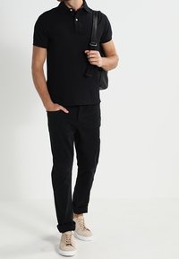 Musta polo-paita, jossa on kaulus, yhdistetty mustiin housuihin ja vaaleanruskeisiin lenkkareihin. Musta olkalaukku on heitetty yhden olkapään yli.