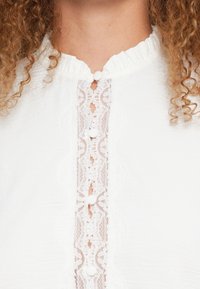 Λευκό μπλουζάκι με στρογγυλεμένο λαιμό, διακοσμητική δαντελένια πλακέτα με κυματιστά άκρα και στρογγυλά κουμπιά από ύφασμα στο κέντρο.